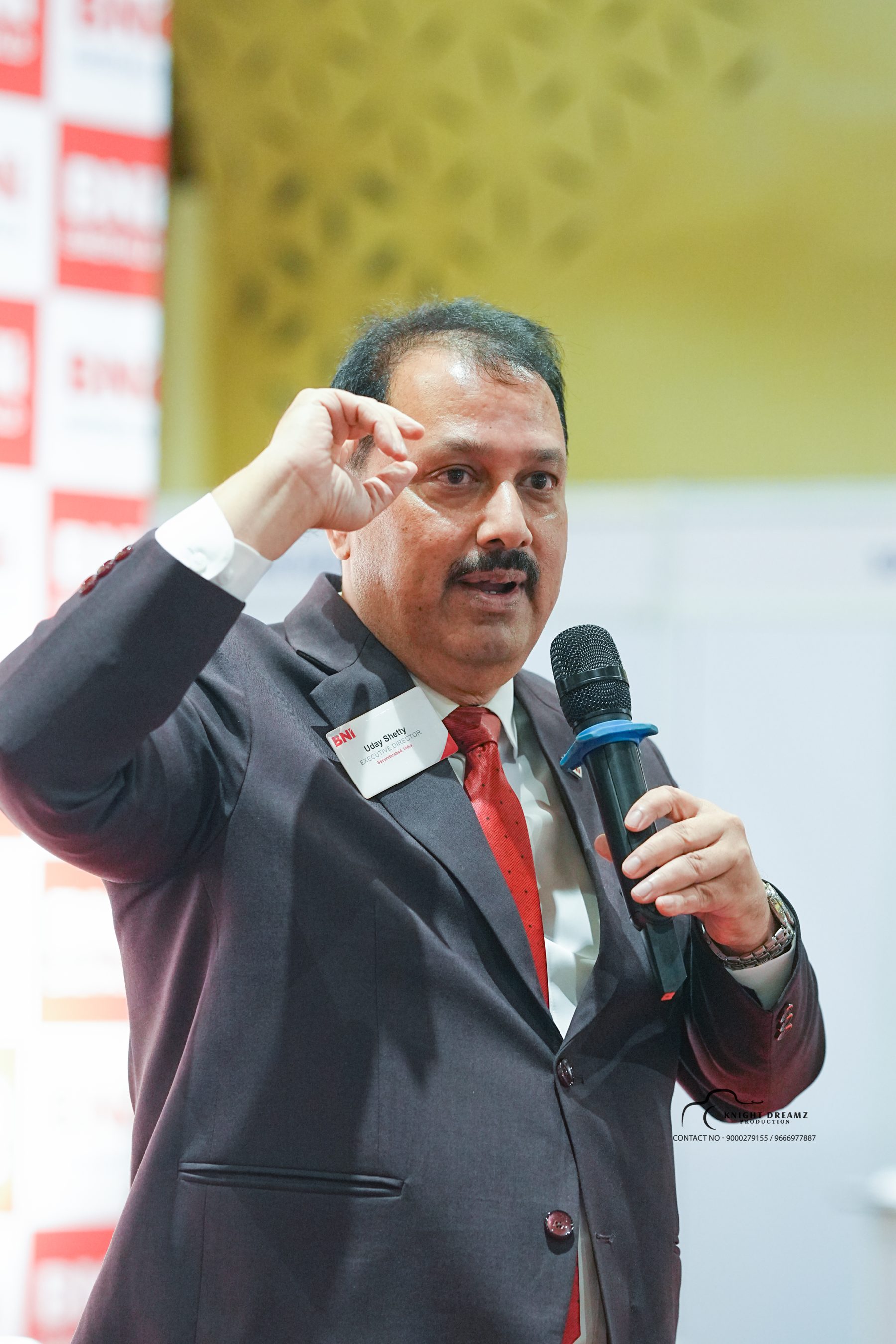 Uday Shetty speaking with microphone — name badge Executive Director